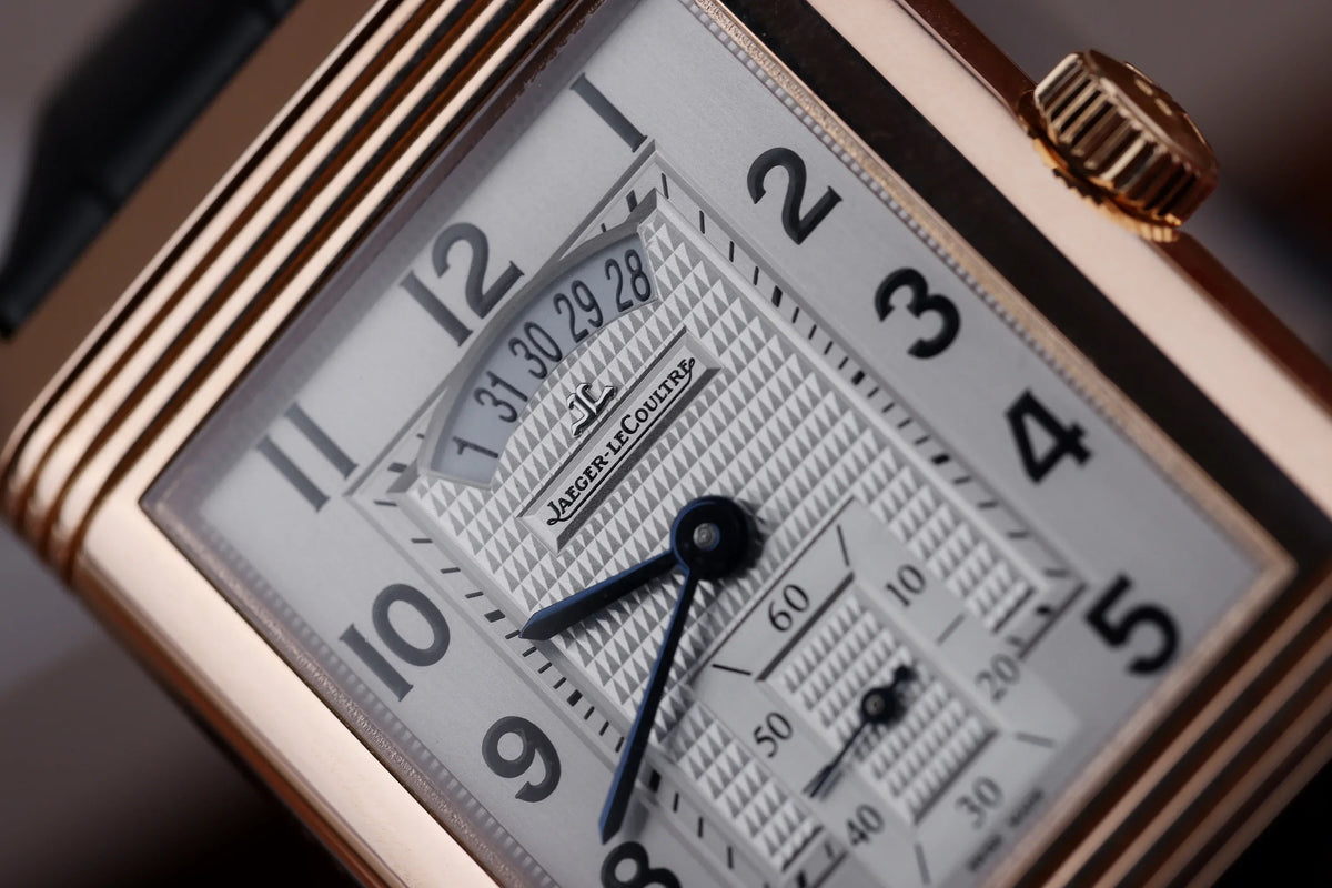 Macro of the silvered guilloche front dial on a Jaeger-LeCoultre Grande Reverso Duo Ref. 273.2.85, showing the arced date aperture below twelve, blued sword hands, small seconds and the reeded rose gold case flank