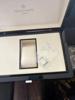 Patek Philippe Geneve original watch box open, showing cream interior with watch cushion and extra strap in plastic wrapping